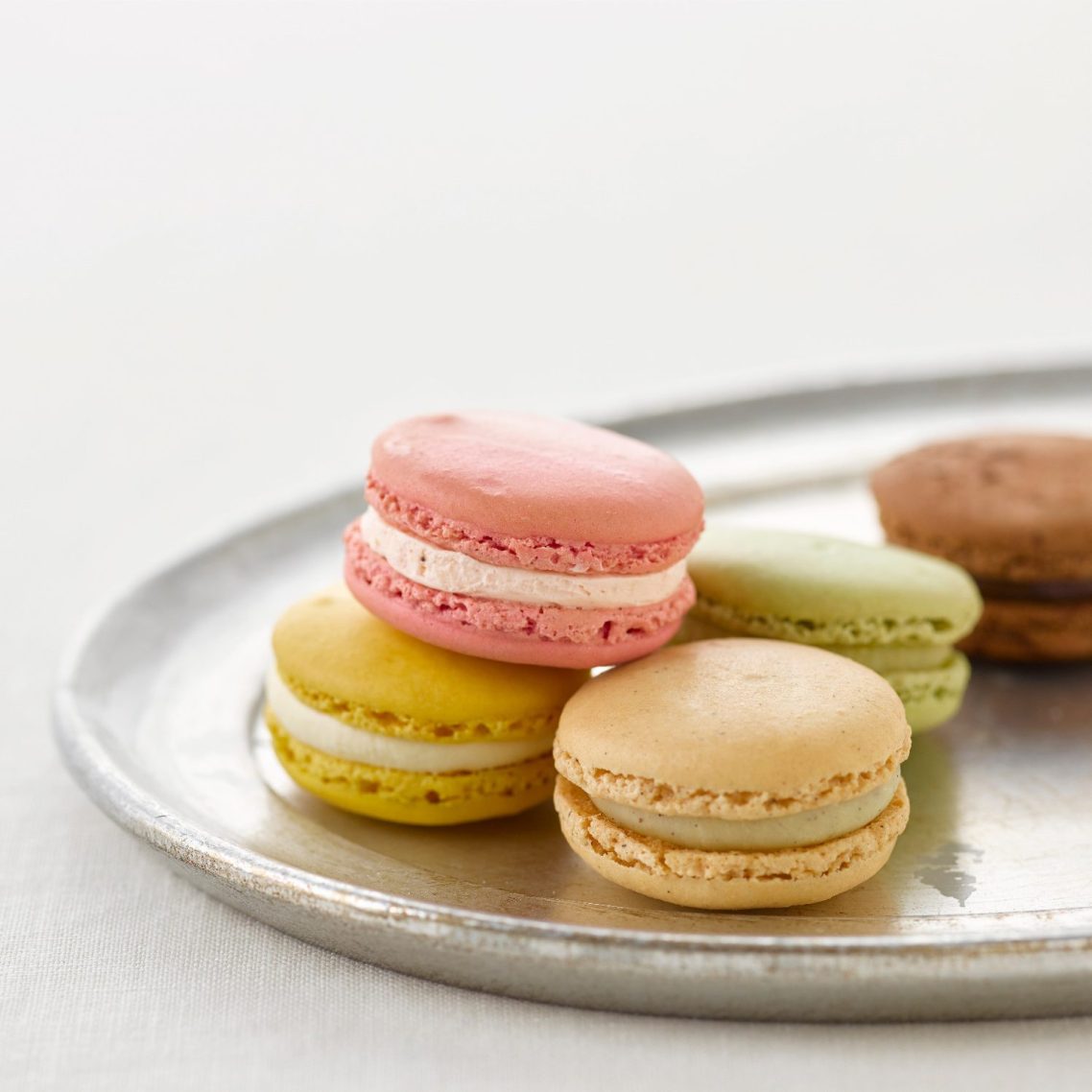Five macarons, one pink, one yellow, on beige, one green, one brown, on a white plate. 