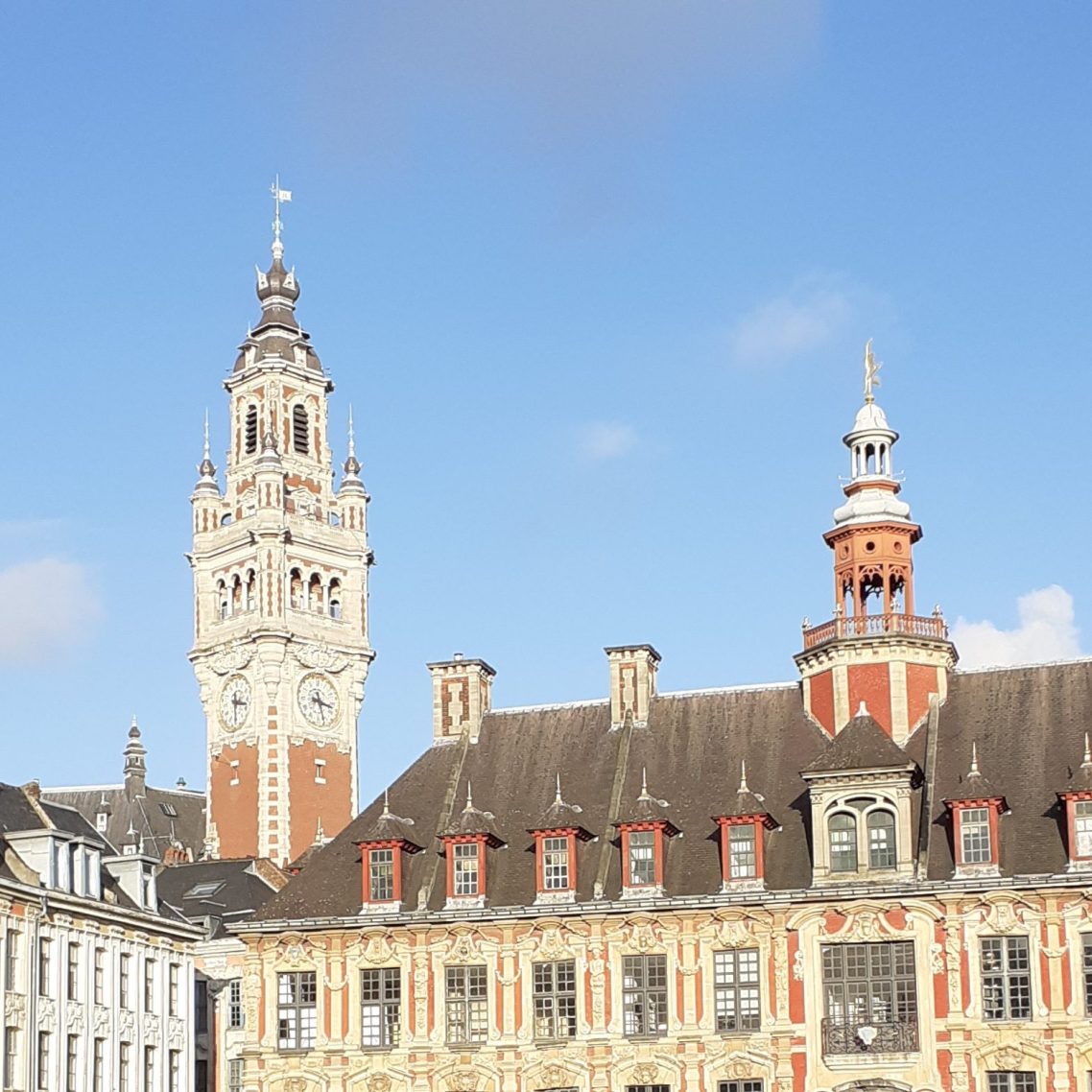 The Beffroi of Lille, a clock tower made of bricks, and the Bourse, a brick building with ornate windows, under a blue sky. 