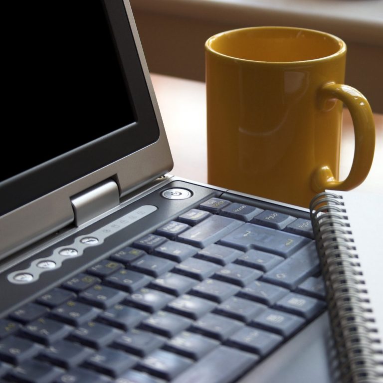 An open laptop, with a yellow mug next to it and a notebook on the keyboard