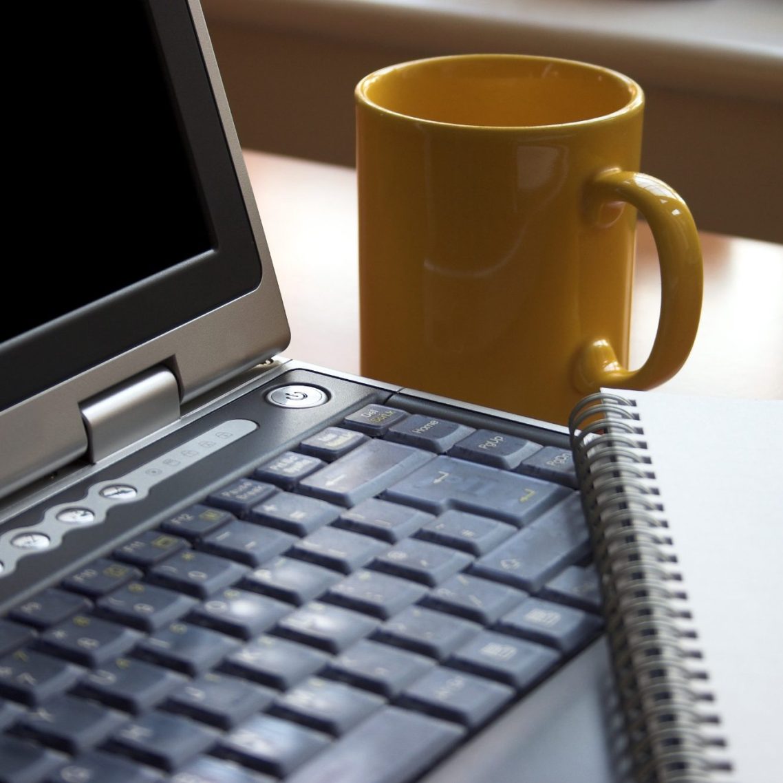 An open laptop, with a yellow mug next to it and a notebook on part of the keyboard. 